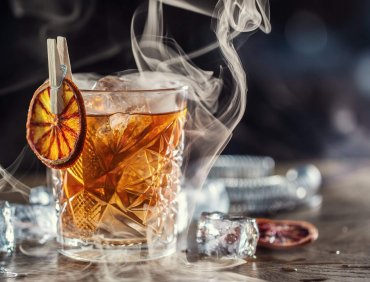 Smoked old fashioned rum cocktail with cubes of ice around on a dark background.