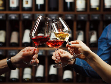 People in good mood clink glasses, celebrating together, photo focus on hands woman and man tasting degustation different types of wine, alcohol beverage to relax, bottles on shelves on background